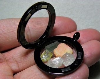 May include: A black locket with a clear glass window, containing small pink, green, and clear gemstones. The locket is open and the gemstones are visible.