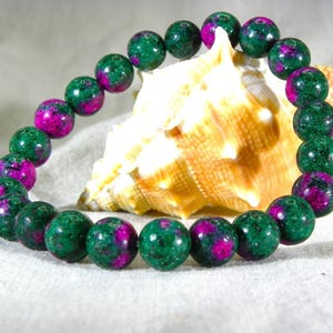 Puede incluir: Pulsera de cuentas redondas y pulidas en tonos verdes y magenta. La pulsera se exhibe frente a una concha marina sobre una superficie blanca. Las cuentas tienen aproximadamente 1,3 cm de diámetro.