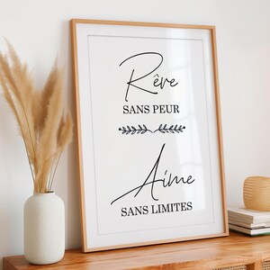 Può includere: Una stampa incorniciata con sfondo bianco e testo nero che recita "Rêve sans peur Aime sans limites". Il testo è in un carattere corsivo ed è circondato da un bordo decorativo di piccole foglie.