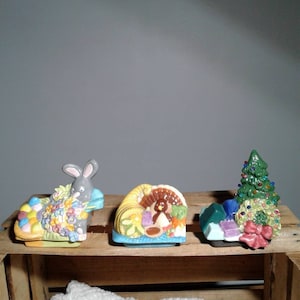 May include: Three ceramic napkin rings, each decorated with a different holiday theme. The first ring features a bunny rabbit surrounded by colorful Easter eggs and flowers. The second ring features a turkey surrounded by fall harvest items. The third ring features a Christmas tree with ornaments and a wreath.