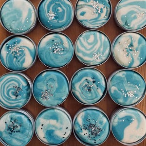 Op de afbeelding: Bovenaanzicht van eenentwintig kleine, ronde, zilveren blikken gevuld met blauw en wit gedraaide deeg. Elk blikje is bedekt met hagelslag. Het deeg heeft een gemarmerd effect, met tinten licht en donkerblauw en wit. De blikken zijn gerangschikt op een houten oppervlak.