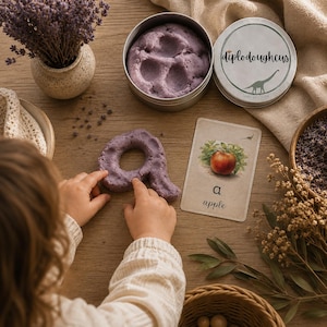 May include: A round metal tin containing purple sensory dough labeled diplodoughcus with a dinosaur graphic, alongside a shaped piece of purple dough and a rectangular card featuring an illustration of an apple with the text a apple below it.