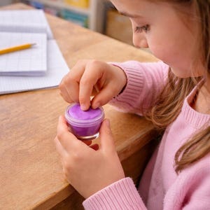 Op de afbeelding: De handen van een kind openen een kleine, doorzichtige pot gevuld met paarse klei. De pot staat op een houten bureau, met een notitieboek en potlood op de achtergrond. Het kind draagt een roze trui.
