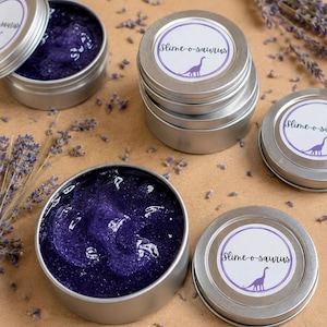Puede incluir: Varios botes pequeños plateados llenos de slime morado, algunos abiertos para mostrar el contenido. Cada bote tiene una etiqueta blanca con el texto "Slime-o-saurus" y una ilustración de dinosaurio morado. Ramas y pétalos de lavanda secos esparcidos.
