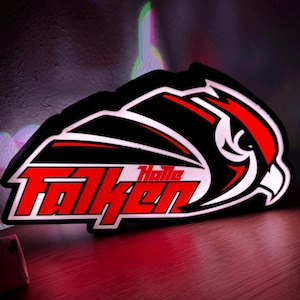 Könnte beinhalten: Ein beleuchtetes Schild in Schwarz, Rot und Weiß mit dem Wort "Falken" in fetter roter Schrift. Das Schild hat die Form eines Vogelkopfes und enthält das Wort "Halle" in kleineren roten Buchstaben.
