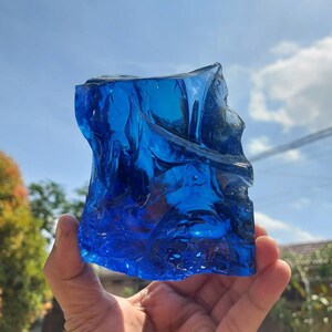 Puede incluir: Un gran trozo de vidrio azul de forma irregular, que se asemeja a un fragmento de hielo, se sostiene en la mano de una persona. El vidrio es transl&uacute;cido y tiene un aspecto ligeramente nublado. El fondo es un cielo azul brillante con algunas nubes blancas.