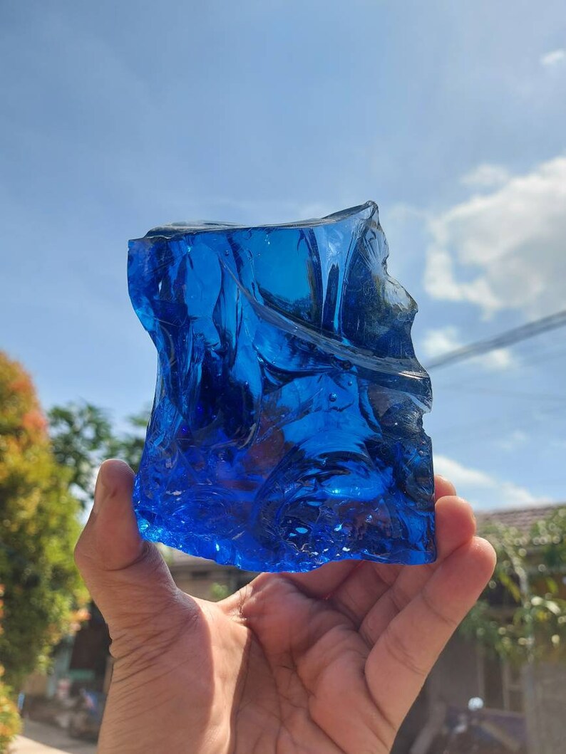 Puede incluir: Un gran trozo de vidrio azul de forma irregular, que se asemeja a un fragmento de hielo, se sostiene en la mano de una persona. El vidrio es transl&uacute;cido y tiene un aspecto ligeramente nublado. El fondo es un cielo azul brillante con algunas nubes blancas.