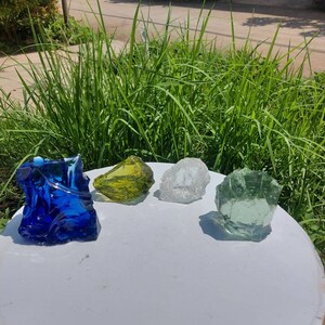 Puede incluir: Cuatro rocas de vidrio en diferentes colores: azul, verde, amarillo y transparente. Las rocas est&aacute;n sobre una superficie blanca.