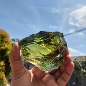 Puede incluir: Una gran pieza de vidrio verde de forma irregular, con una apariencia transl&uacute;cida, casi cristalina. El vidrio se sostiene en la mano de una persona, con un cielo azul y nubes blancas en el fondo.