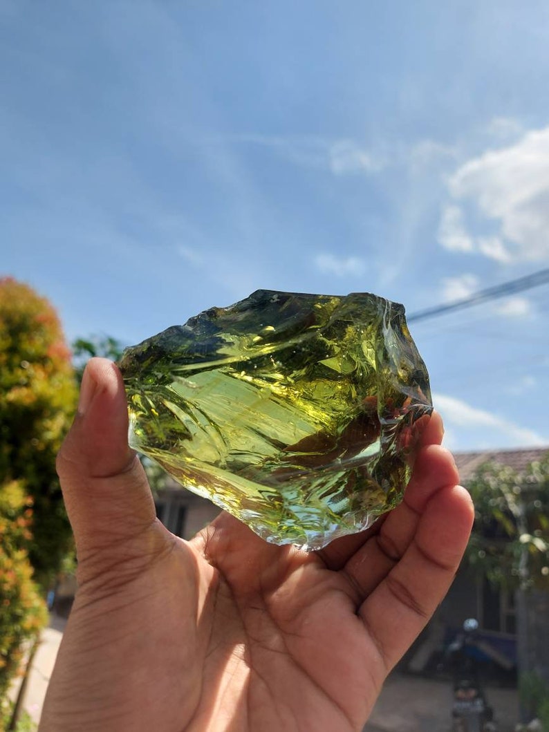 Puede incluir: Una gran pieza de vidrio verde de forma irregular, con una apariencia transl&uacute;cida, casi cristalina. El vidrio se sostiene en la mano de una persona, con un cielo azul y nubes blancas en el fondo.