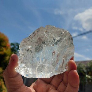 Puede incluir: Un gran cubito de hielo transparente con una superficie rugosa y texturizada. El cubito de hielo se est&aacute; sosteniendo en una mano contra un fondo de cielo azul.