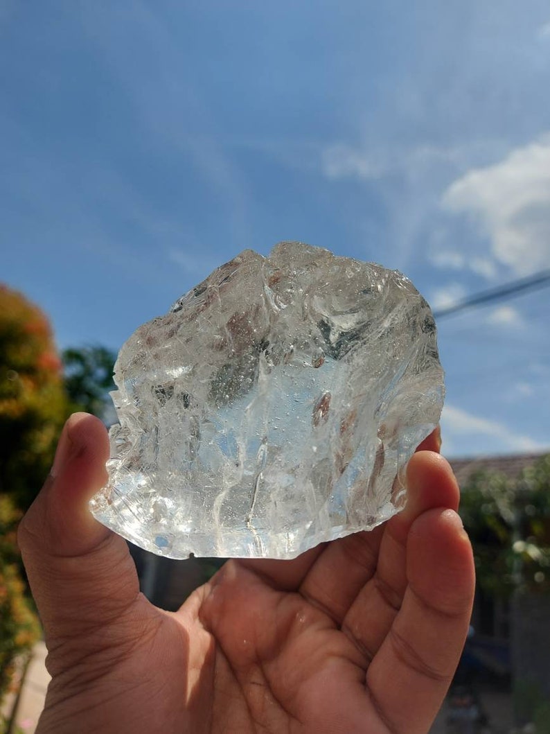 Puede incluir: Un gran cubito de hielo transparente con una superficie rugosa y texturizada. El cubito de hielo se est&aacute; sosteniendo en una mano contra un fondo de cielo azul.