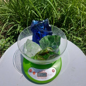Puede incluir: Una b&aacute;scula digital con base verde y pantalla blanca que muestra 17,44 gramos. Un bol de vidrio se encuentra sobre la b&aacute;scula, lleno de trozos de vidrio azul, verde y transparente.