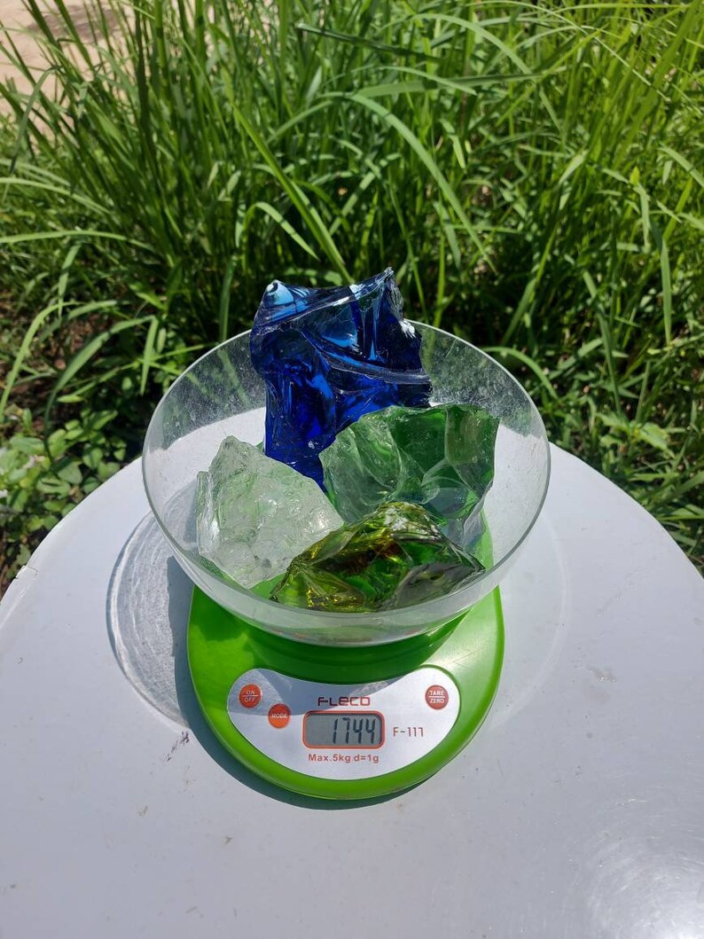 Puede incluir: Una b&aacute;scula digital con base verde y pantalla blanca que muestra 17,44 gramos. Un bol de vidrio se encuentra sobre la b&aacute;scula, lleno de trozos de vidrio azul, verde y transparente.