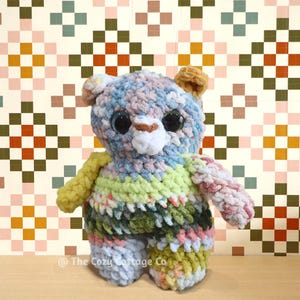 May include: A handmade crochet bear in pastel colors, including blue, pink, green, and yellow. The bear has large, black eyes and a white muzzle. The Cozy Cottage Co. is printed at the bottom of the image.