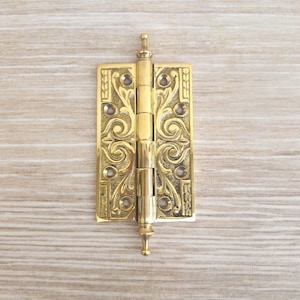 Puede incluir: Una bisagra de puerta de latón pulido con un intrincado patrón de volutas en relieve en ambas placas rectangulares. La bisagra incluye un cilindro central con cinco nudillos y remates decorativos, con seis orificios para tornillos preperforados.