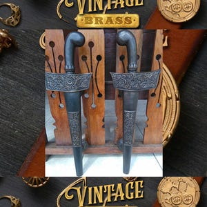 Puede incluir: Dos dagas de metal negro ornamentadas con diseños intrincados se exhiben en un soporte de madera. Las dagas tienen un mango curvo y una hoja puntiaguda. El soporte es de madera oscura con un diseño tallado. Las dagas están etiquetadas como "Vintage Brass".
