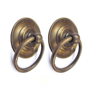 May include: Two antique brass ring pull handles with a round, decorative base. The handles are made of solid brass and have a distressed finish.