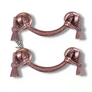 Puede incluir: Dos tiradores de armario de bronce antiguo. Cada tirador presenta un asa decorativa con una barra curvada texturizada y piezas finales detalladas. Los tiradores están sobre un fondo blanco. El texto "VB HARDWARE" es visible.