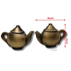 Könnte beinhalten: Zwei schrankknöpfe in Form einer Teekanne in Bronzefarbe. Jeder Knopf ist 4 cm breit und 3 cm hoch. Die Knöpfe haben ein klassisches Teekannen-Design mit Griff, Ausguss und Deckel. Die Knöpfe sind aus Metall und haben einen Vintage-Look.