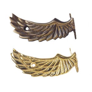 May include: Two decorative metal wing accents, one in a bronze finish and the other in gold. Each wing features detailed feather textures and a small hole for attachment. The wings are likely for furniture or craft projects.