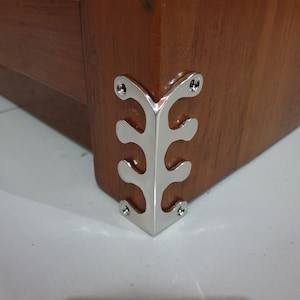 May include: Close-up of a silver corner brace with a decorative design. The brace is attached to a wooden surface.