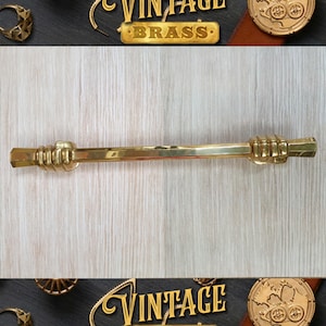 May include: A gold-toned vintage brass door handle with a unique design featuring two hands clasped together. The handle is on a light wood background.