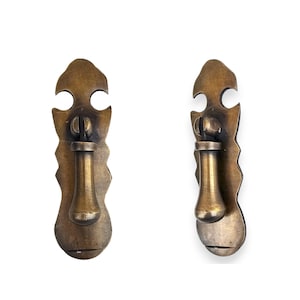 May include: Two matching brass drawer pulls featuring a vertical, elongated backplate with a scalloped edge and a central, downward-hanging drop handle. The metal has a warm, aged patina with visible variations in tone across the surface.