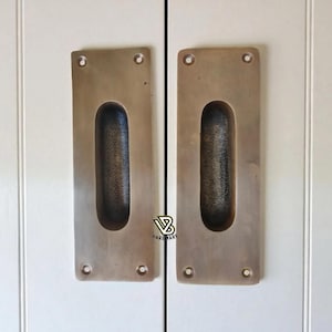 Puede incluir: Dos tiradores rectangulares de armario de color bronce con un agarre ovalado empotrado. Cada tirador tiene cuatro orificios para tornillos y está montado en una puerta de armario blanca. Ferretería.