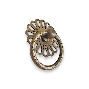 Puede incluir: Tirador de cajón de latón antiguo con placa trasera floral y tirador de anillo. La placa trasera presenta un diseño floral recortado y el tirador de anillo tiene una textura punteada. Este herraje decorativo añade un toque vintage.