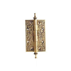 May include: A brass-coloured hinge featuring detailed floral and leaf patterns. The rectangular hinge has rounded edges and decorative finials at the top and bottom. Suitable for doors or cabinets, it measures approximately 10 cm in height.