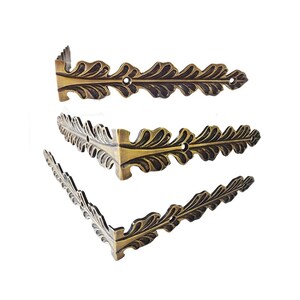 May include: Three L-shaped metal corner brackets with an antique brass finish. Each bracket features a decorative leaf-like pattern along the edges and includes pre-drilled circular holes for mounting purposes, arranged in a vertical stack.