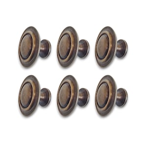 Puede incluir: Un juego de seis pomos circulares para armarios con acabado en bronce oscuro y textura cepillada. Cada pomo presenta una cúpula central elevada y redondeada rodeada por un borde circular plano y ancho, con una base cilíndrica para su montaje.