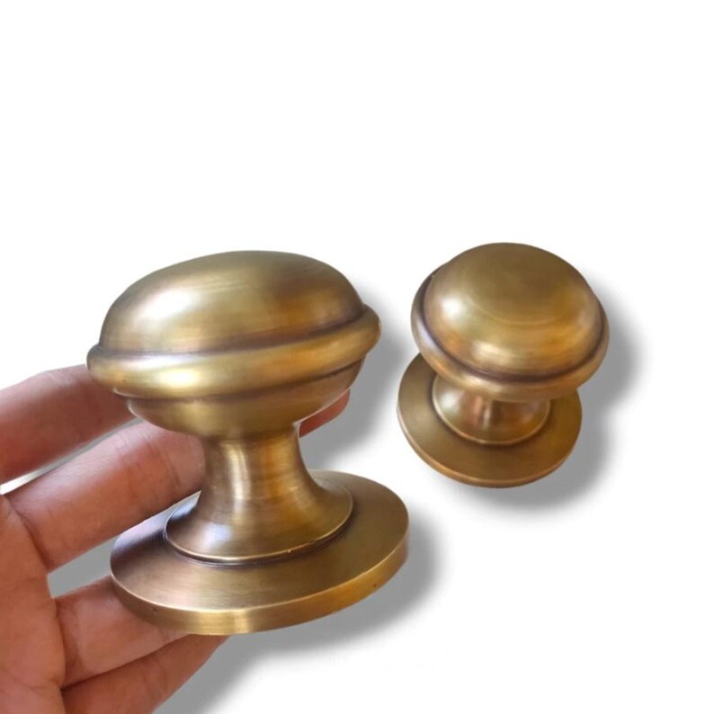 May include: Two antique brass cabinet knobs with a classic design. The knobs have a rounded top and a decorative base. The brass finish gives them a vintage look. One knob is held in a hand, while the other is separate.