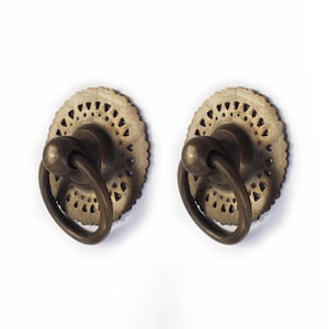 May include: Two antique brass cabinet knobs with a decorative, intricate, circular design. The knobs have a ring pull and are perfect for adding a vintage touch to furniture.