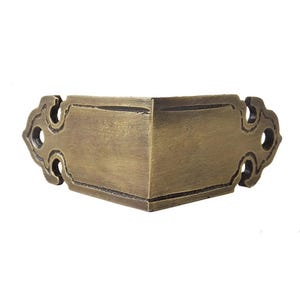 Puede incluir: Un soporte de esquina de color bronce con recortes decorativos. El soporte está diseñado para encajar en una esquina y tiene una forma rectangular con detalles ornamentados. El metal tiene un aspecto ligeramente envejecido.