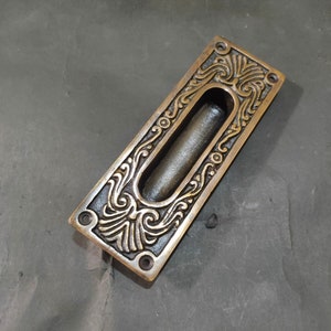 Może przedstawiać: Rektangularny mosiężny klamka do drzwi z ozdobnym wzorem. Klamka ma długą, wąską szczelinę, w którą można włożyć palec.