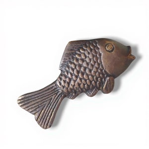 Puede incluir: Un objeto decorativo en forma de pez de color bronce. El pez tiene escamas, aletas y una cola detalladas. El objeto es probablemente de metal y está diseñado como un elemento decorativo.