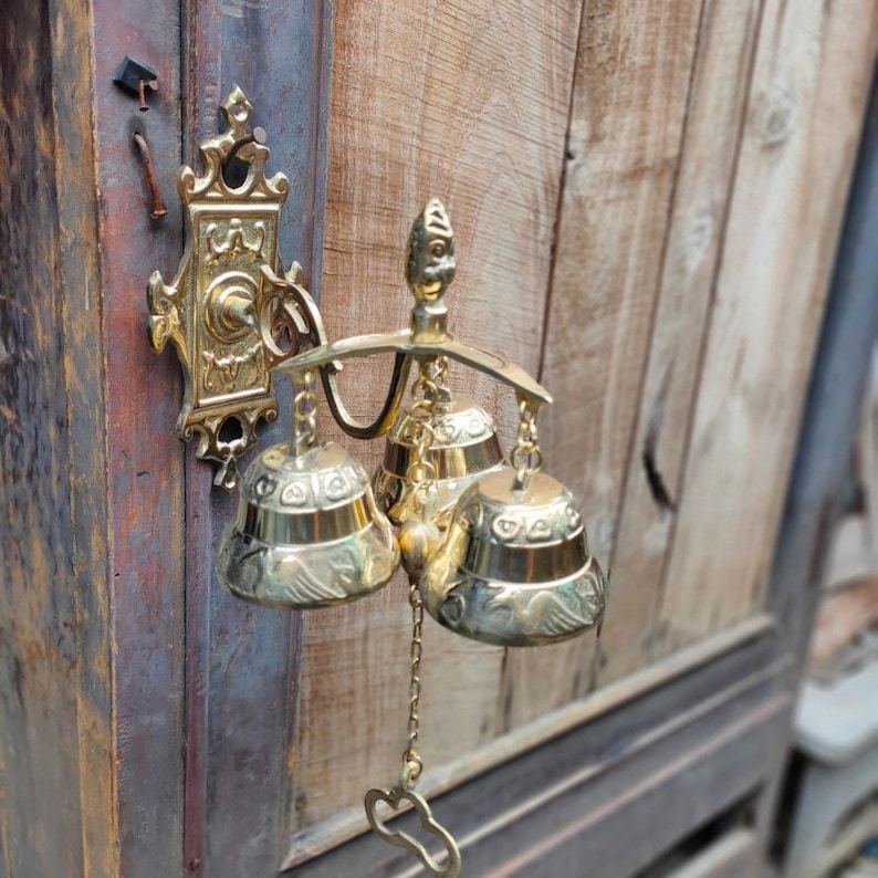 May include: A set of three gold-toned bells hanging from a decorative brass bracket. The bells are adorned with intricate designs and have a traditional style.