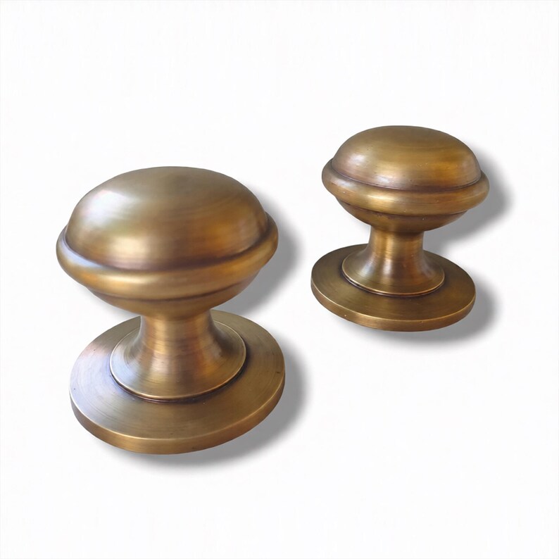 May include: Two antique brass cabinet knobs. Each knob has a rounded top, a circular base, and a cylindrical stem. The knobs have a warm, golden-brown colour and a slightly aged appearance, suggesting a vintage style. They are likely used for furniture or cabinetry.