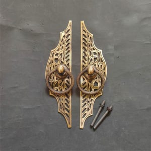 Pode incluir: Duas maçanetas de porta de latão antigo com intrincados desenhos vazados. Cada maçaneta apresenta um puxador de anel decorativo e é acompanhada por dois parafusos. As maçanetas têm um acabamento quente e envelhecido, sugerindo um estilo vintage ou artesanal.