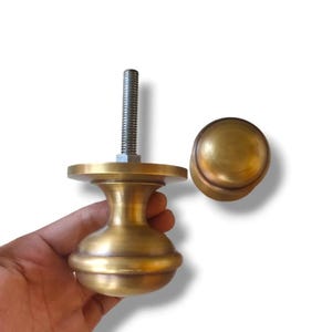 May include: Two brass cabinet knobs. One has a threaded rod and a nut, the other is a simple round knob. The knobs are a warm gold colour and appear to be made of metal. The knobs are on a white background.