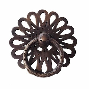 May include: A decorative brass door pull with a circular, ornate design. The pull features a ring handle and a detailed, floral-like pattern.