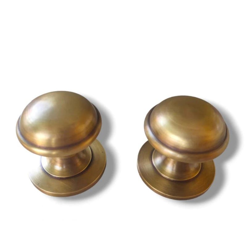 May include: Two antique brass cabinet knobs. Each knob has a rounded top and a circular base. The knobs have a warm, golden hue and are likely made of solid brass. They are designed for easy installation on cabinet doors or drawers.