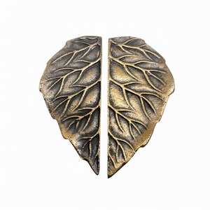 May include: A pair of matching metal cabinet pulls shaped like two halves of a single leaf. The hardware features a dark, antique brass finish with a textured, raised vein pattern across the surface, creating a detailed botanical appearance.