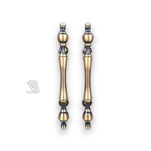 Może przedstawiać: Dwa uchwyty do szafek w stylu antycznym z brązowym wykończeniem. Każdy uchwyt ma dekoracyjny design z centralnym, wydłużonym uchwytem i ozdobnymi końcami. Uchwyty są symetryczne i przeznaczone do okuć meblowych.