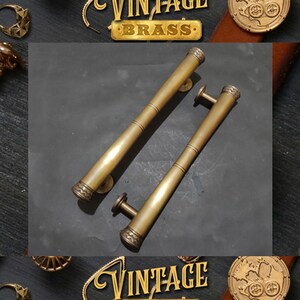 Op de afbeelding: Een paar vintage messing deurgrepen met een decoratief ontwerp. De grepen zijn lang en cilindrisch met een afgerond uiteinde. De grepen zijn gemaakt van massief messing en hebben een gepolijste afwerking.