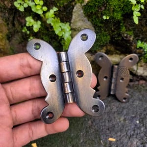 May include: Two antique bronze-coloured butterfly-shaped hinges. Each hinge has a decorative butterfly design with rounded edges and multiple screw holes. The hinges are made of metal, likely for furniture or decorative use.