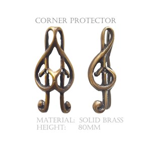 May include: Two solid brass corner protectors shaped like musical treble clefs. The text CORNER PROTECTOR, MATERIAL: SOLID BRASS, and HEIGHT: 80MM is printed on the image. The metal has a dark, antique-toned finish with visible curves and openings.