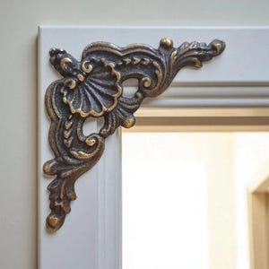 May include: Ornate bronze-colored corner bracket with a detailed, symmetrical design. The bracket features a central fan-like element and scrolling leaf patterns. It is mounted on a white frame, suggesting a decorative or architectural element.